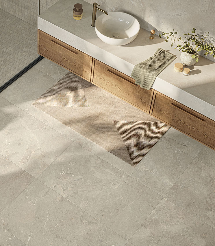 Bagno contemporaneo con superfici delle collezioni Revel Dune, Mosaico Dune e Workshop Jade, caratterizzato da pavimento e rivestimenti effetto pietra nei toni chiari. L’ambiente presenta un lungo mobile lavabo in legno, piano chiaro con lavabo da appoggio bianco, rubinetteria dorata, tappeto geometrico e doccia con mosaico coordinato.