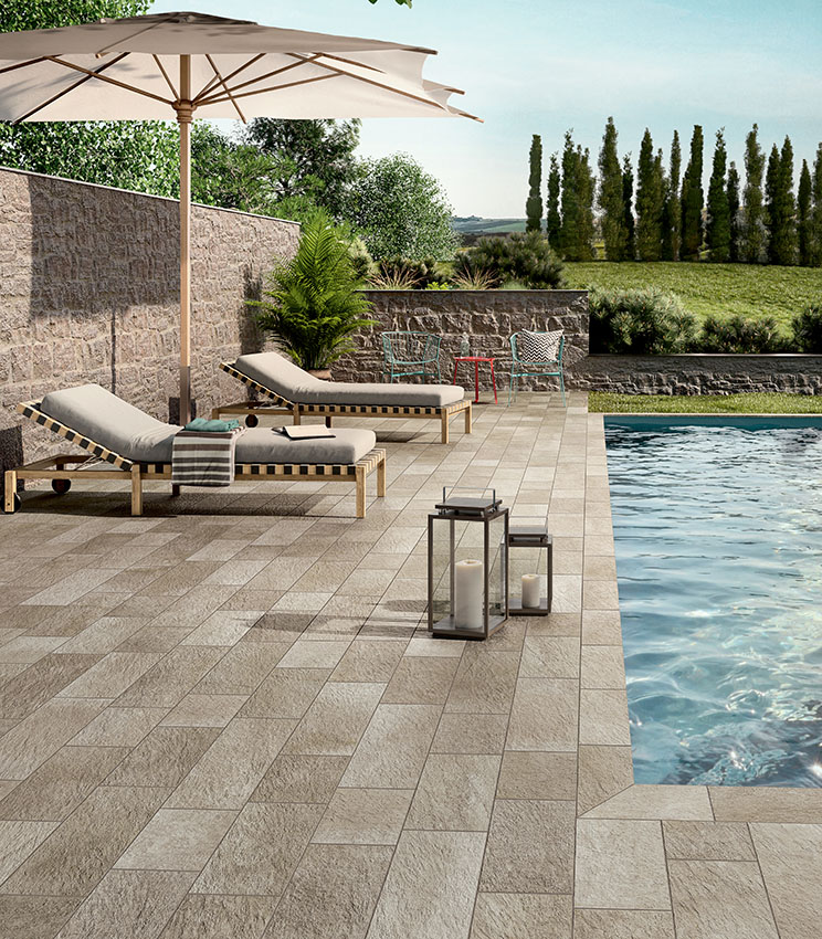 Area piscina con pavimentazione in gres porcellanato effetto pietra, lettini prendisole e arredi outdoor, per uno spazio elegante, naturale e resistente.