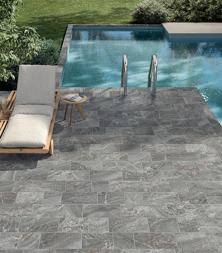 Area piscina con pavimentazione in gres porcellanato effetto pietra grigia, lettino prendisole e scala d’accesso, per uno spazio elegante, naturale e resistente.